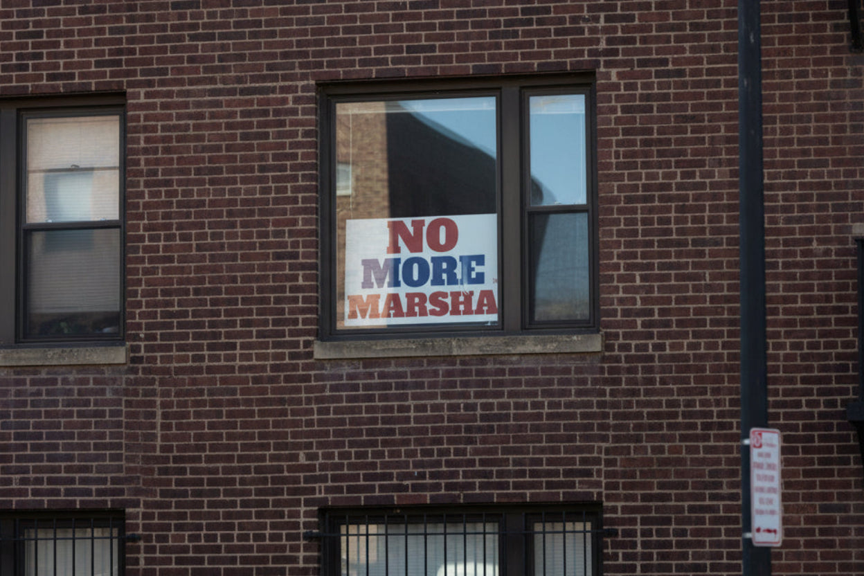 Political sign with 'NO MORE MARSHA' text in red and blue on a white background placed in the window of a brick apartment building.