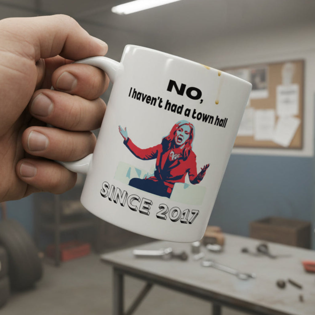 White mug with a graphic of Marsha Blackburn and black text 'No, I haven't had a town hall since 2017' on a white background, with a white man holding it in a mechanic's shop